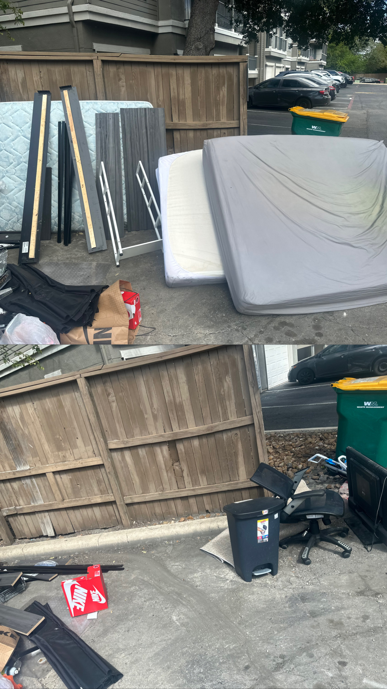 Discarded mattresses, bed frame parts, chairs, trash bin, and other household items piled near a wooden fence in a parking lot.