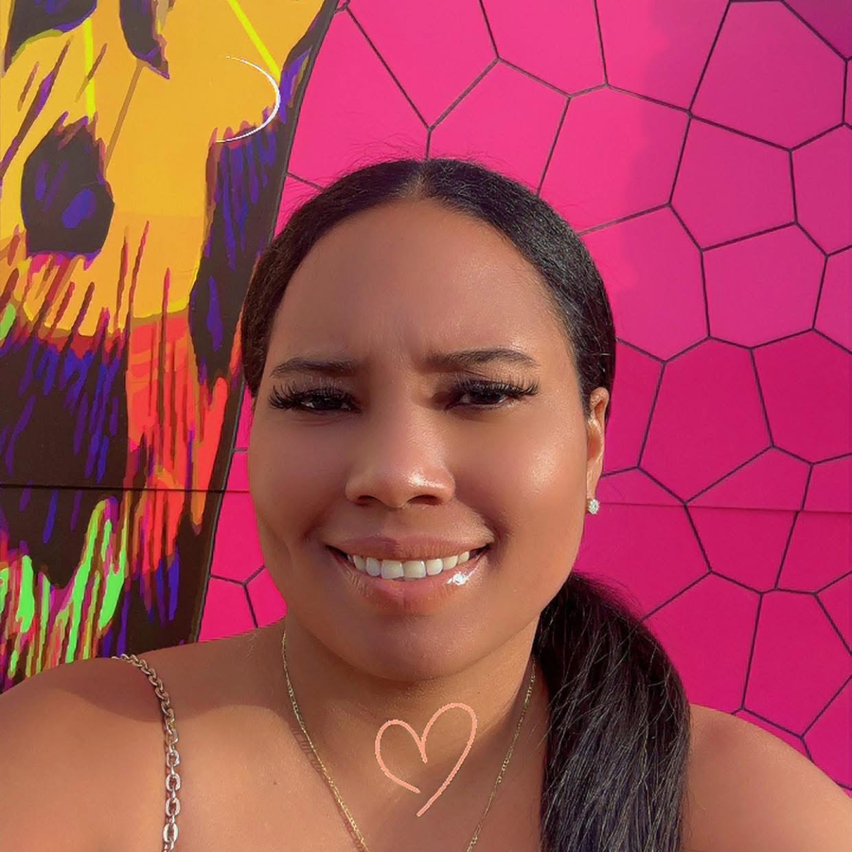 Close-up of a smiling woman with long dark hair and a heart drawn on her chest, standing in front of a bright pink geometric wall and colorful mural.